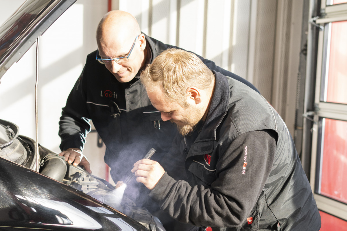 Zwei Mechatroniker untersuchen mit einer Taschenlampe den Motorraum eines Autos